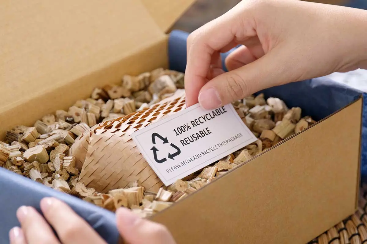 Hand placing a recyclable label on shredded kraft paper packaging inside a cardboard box, promoting sustainable shipping.